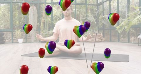 Man meditating on yoga mat in sunroom conservatory with floating rainbow heart balloons