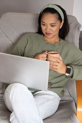 Asian woman relaxing and working on laptop while holding coffee in cozy armchair at home