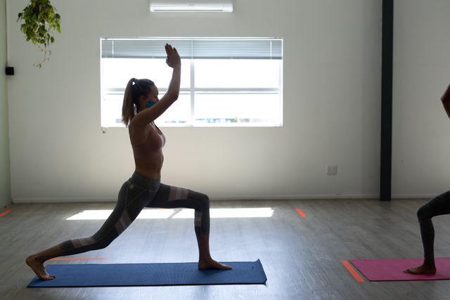 Caucasian woman on a yoga mat stretching doing yoga while wearing a facemask. infront of her is another woman doing yoga,