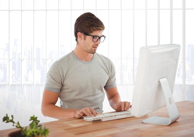 Young man in spectacles working on desktop computer in a modern office environment. Ideal for use in business, technology, and professional workspace contexts. Can be used for promoting office productivity, tech products, or business services.