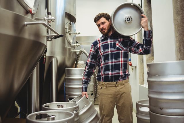 Young man carrying kegs in a brewery, surrounded by brewing equipment. Ideal for use in articles or advertisements related to the craft beer industry, brewery operations, or industrial manufacturing. Can also be used for promoting brewery tours or showcasing the brewing process.
