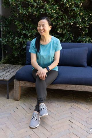 Mature asian woman sitting smiling on blue terrace sofa wearing smartwatch activewear