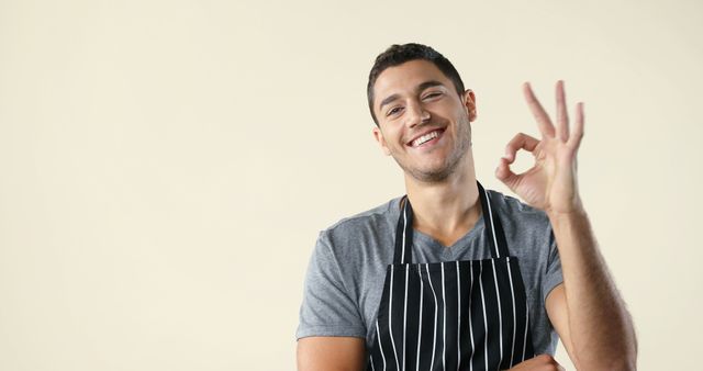 Young man wearing an apron smiling and making an OK hand gesture against a plain background. Ideal for use in culinary blogs, cooking classes promotions, restaurant advertisements, or positive encouragement messages.