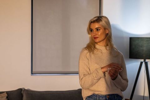 Young woman in beige sweater standing by sofa, warm cozy living room lifestyle portrait