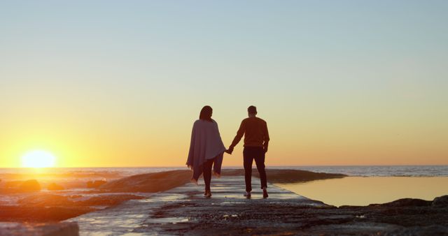 A young couple walks hand in hand along a pier at sunset, with copy space. Their silhouette against the vibrant sky creates a romantic and peaceful atmosphere.