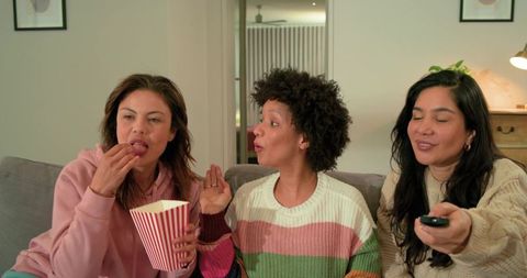 Friends enjoying cozy movie night on couch eating popcorn using remote for home entertainment