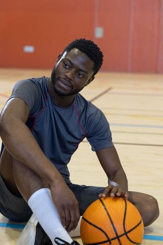 Contemplative african american basketball player sitting on hardwood court holding ball