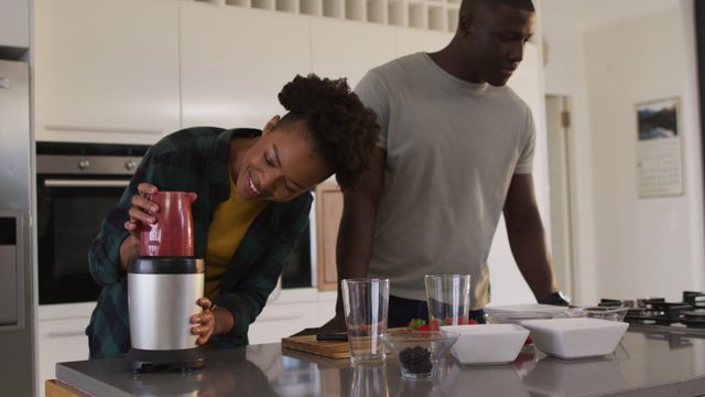 African American couple happily preparing fresh juice using a blender in a modern kitchen. Scene radiates love, joy, and a healthy lifestyle. Perfect for use in advertisements, blog posts, or social media promoting family togetherness, kitchen appliances, health and wellness, or modern lifestyles.