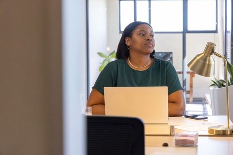 African american woman working at laptop in bright modern coworking office, looking thoughtful