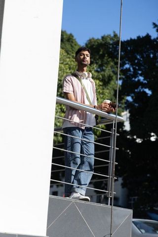 Indian man standing on urban terrace holding coffee and smartphone wearing headphones