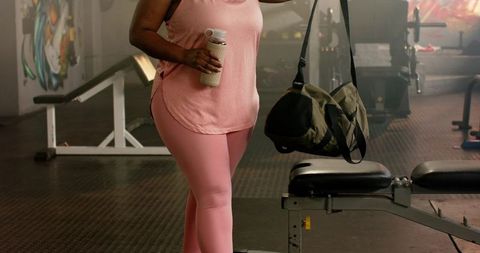 African american woman standing in gym holding protein shaker with camo duffel bag