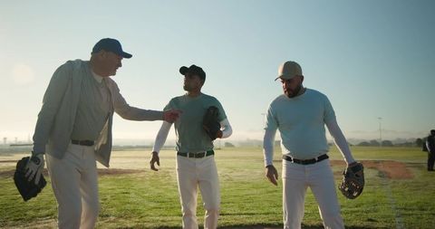 Senior Baseball Coach Walking with Teammates on Dewy Field at Sunrise Showing Teamwork