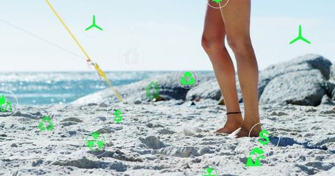 Woman standing barefoot on sandy beach showing ankle bracelet with green eco icons