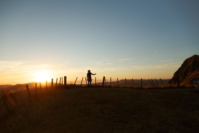 An individual standing near a fence on a hill during sunset, with the sky painted in warm colors. Suitable for use in travel blogs, adventure articles, nature websites, and outdoor lifestyle promotions. Ideal for illustrating themes of tranquility, exploration, solitude, and connection with nature.