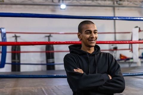 Confident young man leaning on boxing ring ropes wearing black hoodie gym training