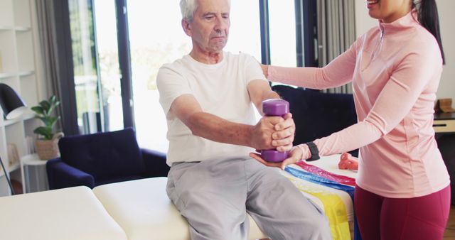 Senior man performing physical therapy exercises with the guidance of a female therapist in a home setting. Man lifting purple dumbbells while the therapist assists and instructs him. This image highlights themes of senior health, rehabilitation, and physical fitness. Useful for promoting healthcare services, rehabilitation centers, senior wellness, and fitness programs.