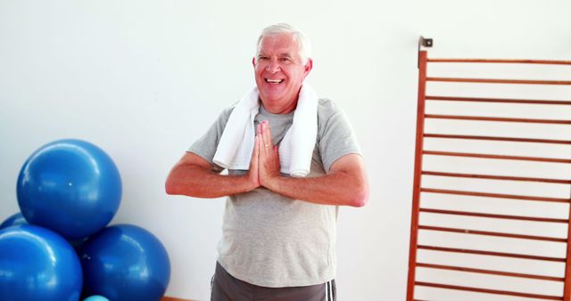 Senior man is practicing yoga at a fitness studio with a towel around his neck. He is smiling and looks relaxed, standing near some blue exercise balls and wooden fitness equipment. This image can be used for promoting wellness programs, senior fitness classes, healthy lifestyle, and yoga or exercise routines for the elderly.