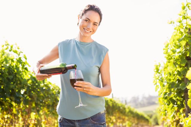 Woman standing in vineyard on sunny day, pouring red wine from bottle into glass. Ideal for use in articles about wine tasting, vineyard tours, wine production, outdoor leisure activities, and lifestyle blogs.