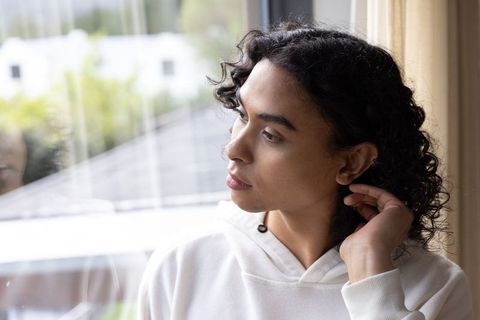 Non-binary adult gazing through window touching hair in white hoodie, reflective indoor portrait