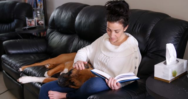 Indoor scene of woman reading book while sitting with her dog on a couch. Suitable for topics on home relaxation, leisure activities, reading habits, pet companionship, and domestic lifestyle. Ideal for articles or advertisements focused on home comfort, pet products, or wellness.