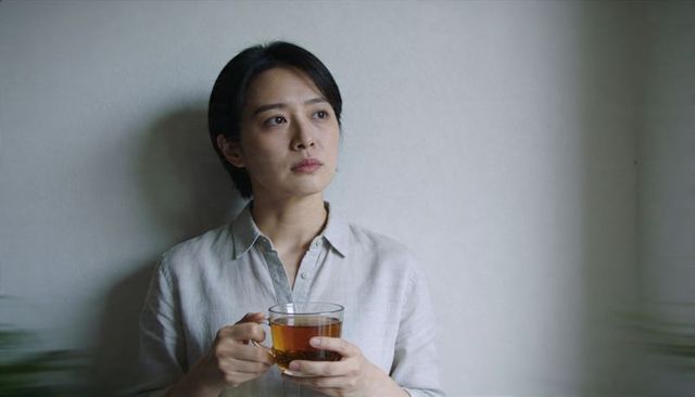 Contemplative woman holding glass mug of tea by pale gray wall, minimalist interior