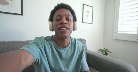 Young african american man wearing headphones taking selfie on couch in minimal living room