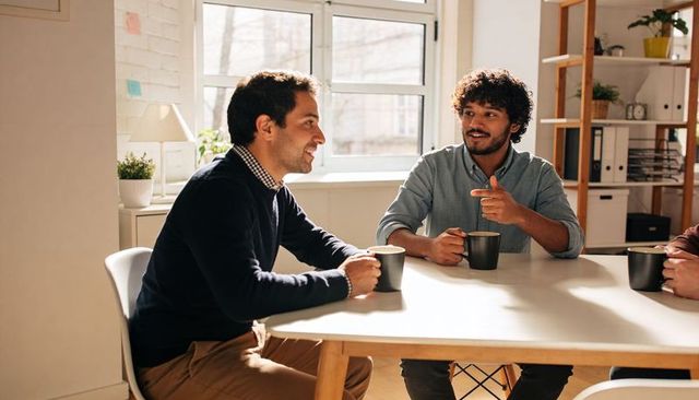Colleagues having coffee and conversing in bright modern office during casual meeting