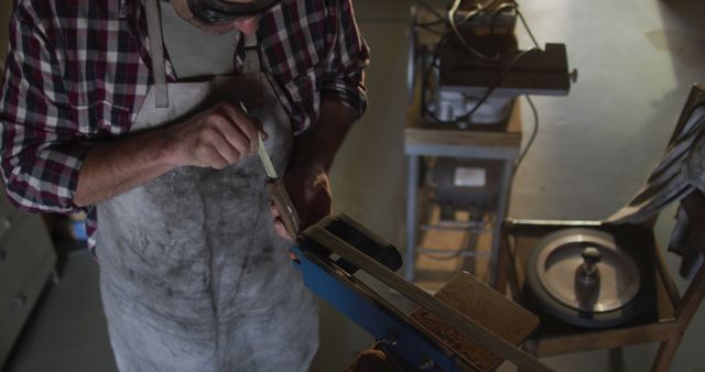 Craftsman sharpening chisel in workshop using power tool while wearing apron and plaid shirt. Detail shows hands holding the chisel against grinder. Great for illustrating woodworking, craftsmanship, DIY projects, and tools. Suitable for blogs, magazines, educational materials, promotional content for woodworking classes or tool brands.
