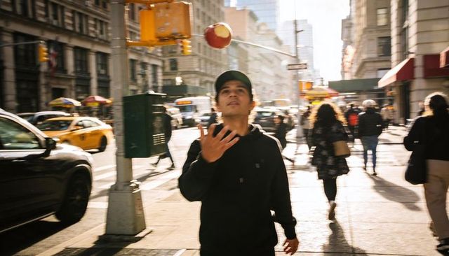 Sunlit urban street with young man tossing red ball, hoodie and baseball cap focus