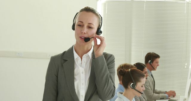 A professional Caucasian woman in business attire is working as a customer service representative, with copy space. She is using a headset to communicate, while her diverse colleagues are focused on their calls in the background.
