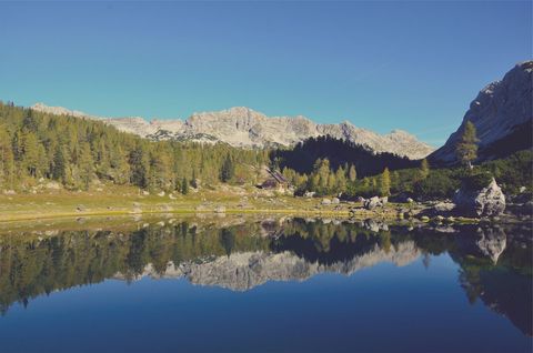 Mountain Lake Reflecting Alpine Cabin, Pine Forest and Rugged Limestone Peaks under Clear Blue Sky