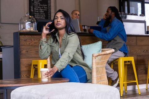 Indian woman talking on smartphone while reaching for takeaway coffee cup in cozy cafe lounge