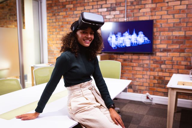 Portrait of a confident happy biracial businesswoman with long dark hair working in a modern architects creative office, wearing a Virtual Reality headset, sitting on a desk, looking at camera, brick wall in the background.