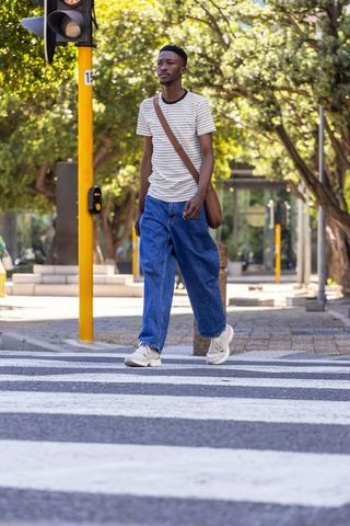 African american man walking across zebra crosswalk under tree canopy on urban street