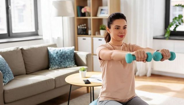 Young woman exercising on stability ball with dumbbells in bright modern living room