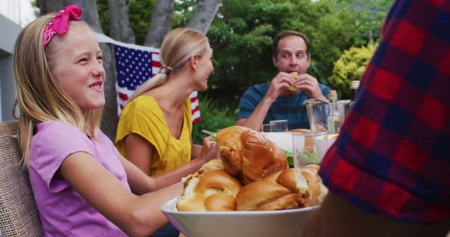 The scene depicts a family enjoying a meal together outside. A girl in a purple shirt and a woman in a yellow shirt are smiling, while a man in a blue shirt is eating a burger. An American flag is visible in the background, suggesting a possible holiday context like Independence Day. This image can be used in advertisements, blog posts, or articles about family, food, holidays, and summer activities.