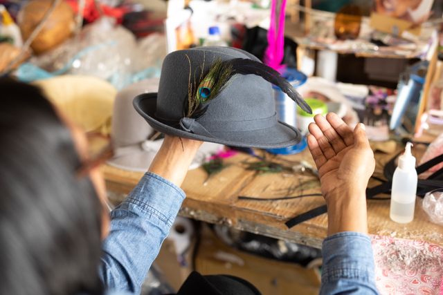 Woman decorating hat with feather in workshop, showcasing craftsmanship and creativity. Ideal for use in articles about handmade fashion, artisan crafts, or creative workspaces. Suitable for promoting hat-making workshops, fashion design courses, or DIY craft projects.