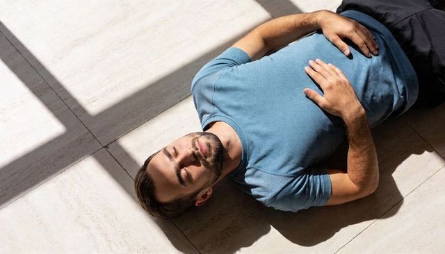 Young man lying on sunlit tiled floor with window-frame shadow, calm minimalist interior relaxation