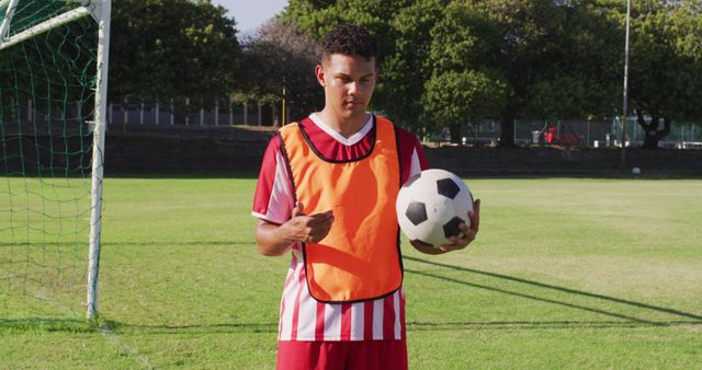 A teenage soccer player in a red and white uniform is standing on a soccer field under bright daylight, holding a soccer ball. Trees can be seen in the background, providing a natural environment for practice. This image is perfect for sports-related articles, youth sports promotion, educational materials on soccer techniques, or advertising for sports equipment and training programs.