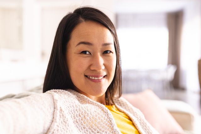 Asian woman sitting comfortably in a bright living room, smiling and relaxed. Ideal for use in lifestyle blogs, articles on home comfort, mental health, inclusivity, and domestic life. Perfect for promoting relaxation and well-being.