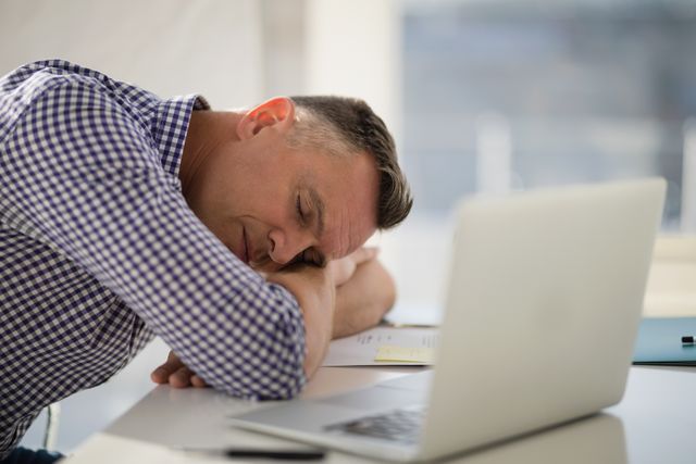 Middle-aged male executive in checkered shirt resting head on arms at desk with laptop. Ideal for illustrating workplace fatigue, stress, and the importance of breaks. Suitable for articles on work-life balance, corporate wellness, and productivity.