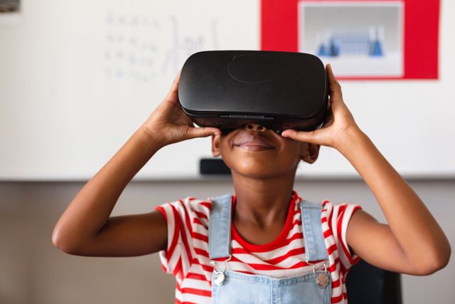 Smiling african american elementary schoolgirl wearing vr glasses while sitting at desk in classroom. unaltered, virtual reality simulator, playful, fun, technology and school concept.