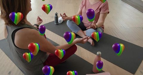 Two women practicing seated yoga meditation with floating rainbow hearts for mindful pride