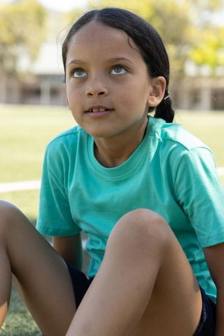 Preteen girl sitting on grass wearing teal shirt and dark shorts gazing upward in park
