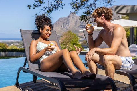 Mixed race couple relaxing poolside sipping cocktails on sunny terrace with ocean view