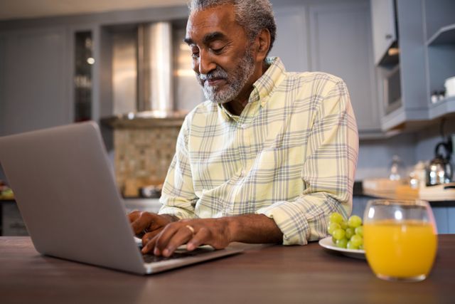 Senior man using laptop in kitchen, smiling while working or browsing internet. Ideal for concepts of technology use among elderly, home office, modern lifestyle, and healthy living.