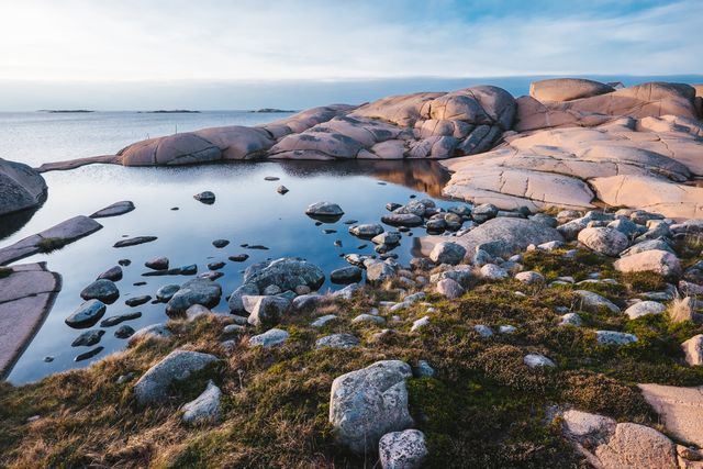 This image captures a serene coastal landscape featuring rocky formations by calm waters at dusk. Knolls and boulders dominate the foreground, with a glassy sea stretching towards the horizon under a waning twilight sky. Perfect for use in travel brochures, nature blogs, wallpapers, and promotional materials emphasizing peace, tranquility, and natural beauty.