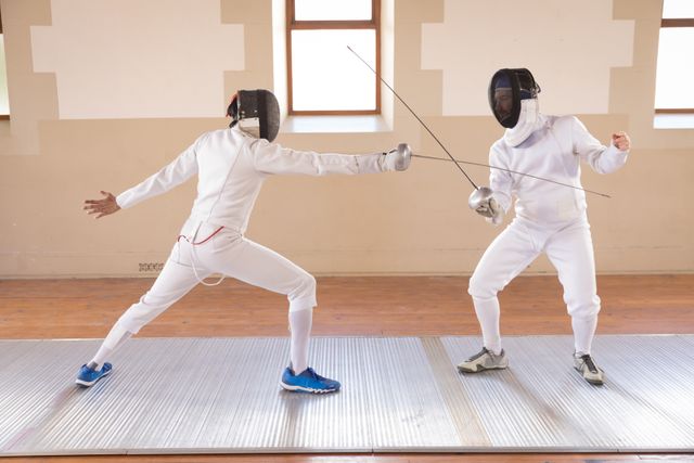 Two people in a fencing duel, wearing protective fencing clothes, holding sabers in a gym. Sport and working out.
