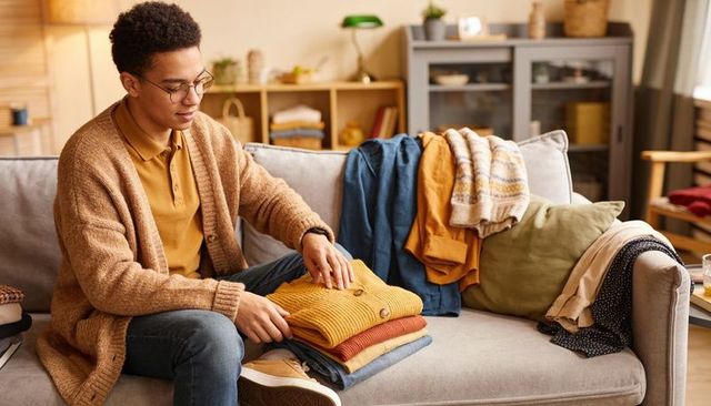 Young man folding knit sweaters on sofa for cozy autumn wardrobe organization and home styling
