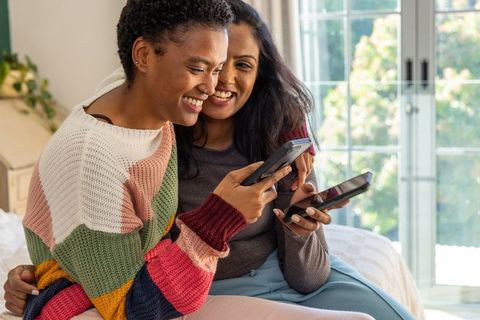 Smiling black and south asian women sharing smartphones on sunlit sofa, cozy home lifestyle